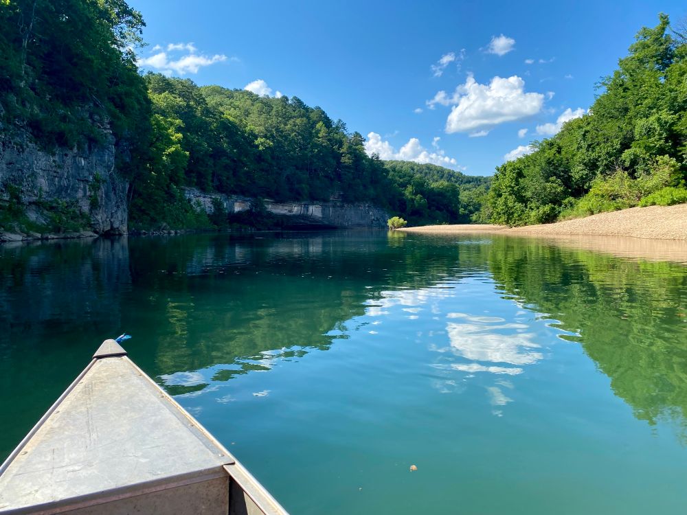 A canoe floating down a river with a blue partly cloudy sky overhead and green trees on the bank and bluffs.