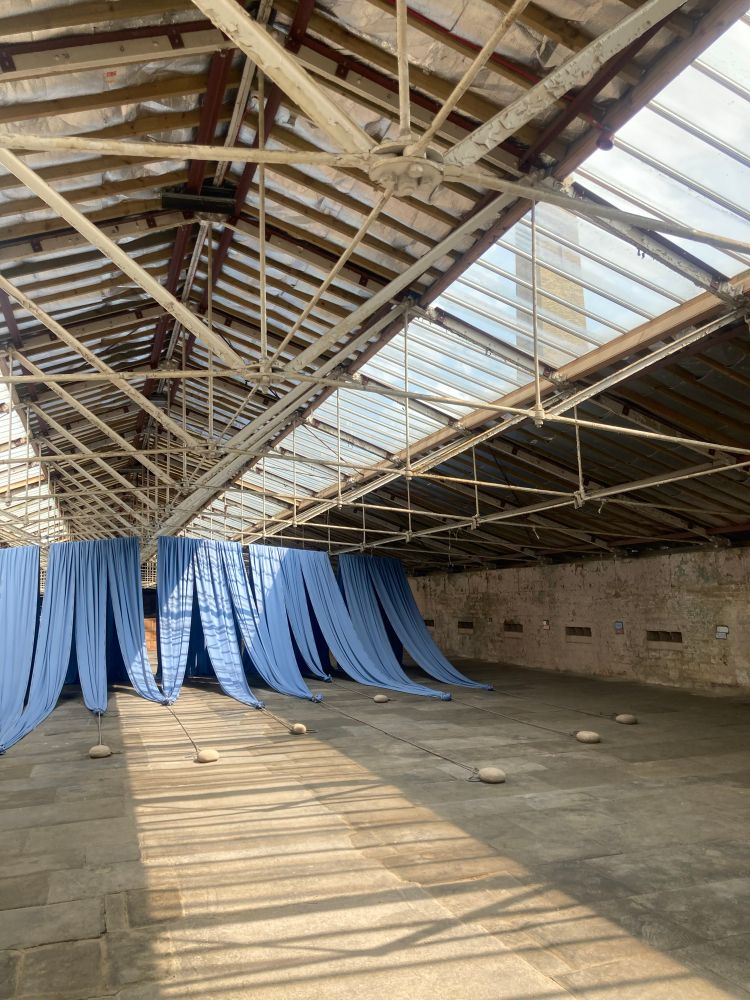 Yards and yards of cornflower blue material is draped at one end of a large industrial room with glass windows in the roof and a factory chimney in the distance. 