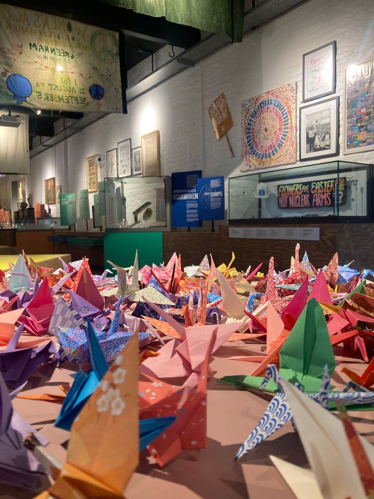 A great many origami birds of all colours and patterns in the foreground, with exhibition displays about peace in the background. 