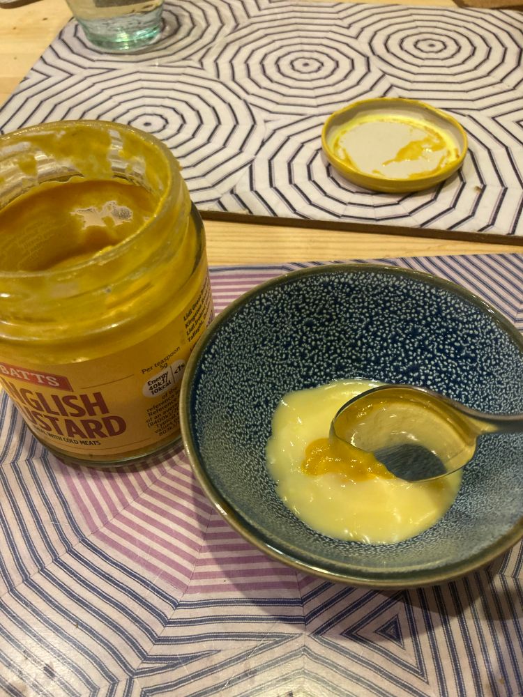 It’s a small bowl of light yellow custard with a teaspoon of mustard in it, the offending mustard jar sits to the left.