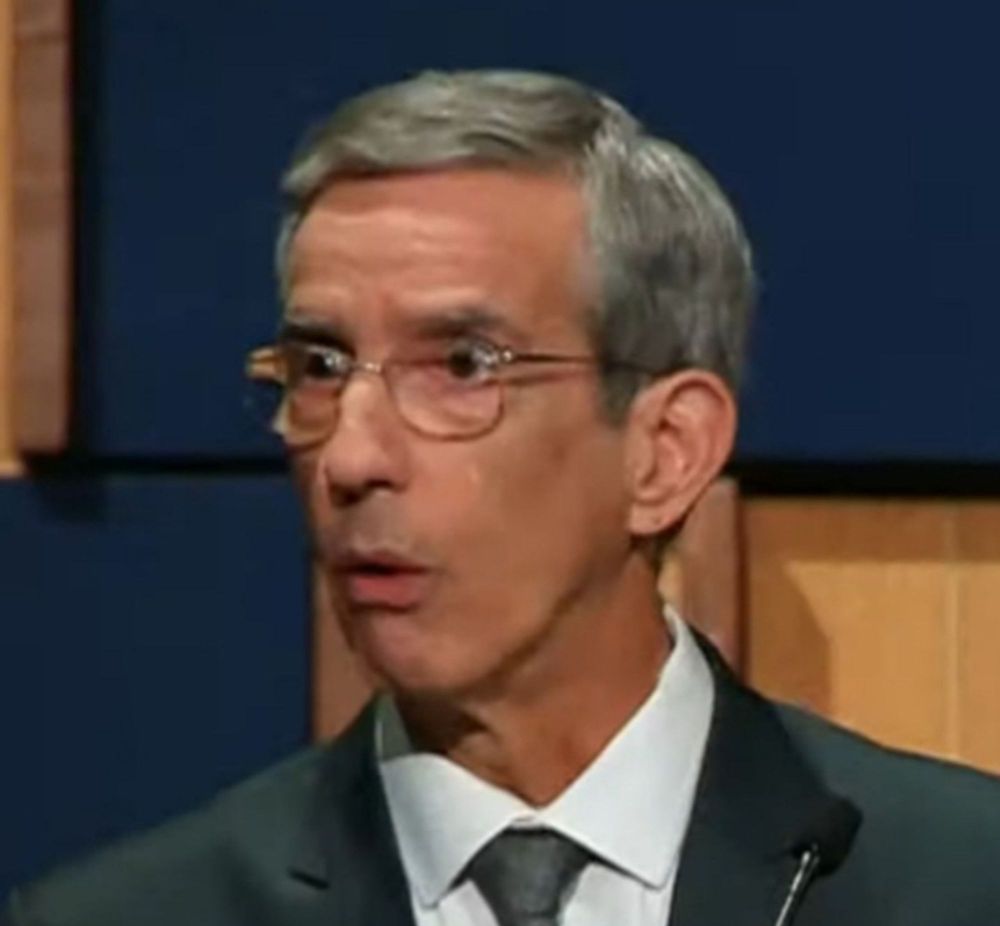 O jornalista Leão Serva, um homem branco de 65 anos, cabelos grisalhos curtos e óculos de armação fina, trajando um paletó escuro com gravata cinza-clara e camisa branca. Sua expressão é de surpresa e perplexidade, com olhos arregalados e a boca ligeiramente aberta, enquanto assiste, fora de quadro, o candidato Datena agredir o candidato Pablo Marçal com uma cadeirada.