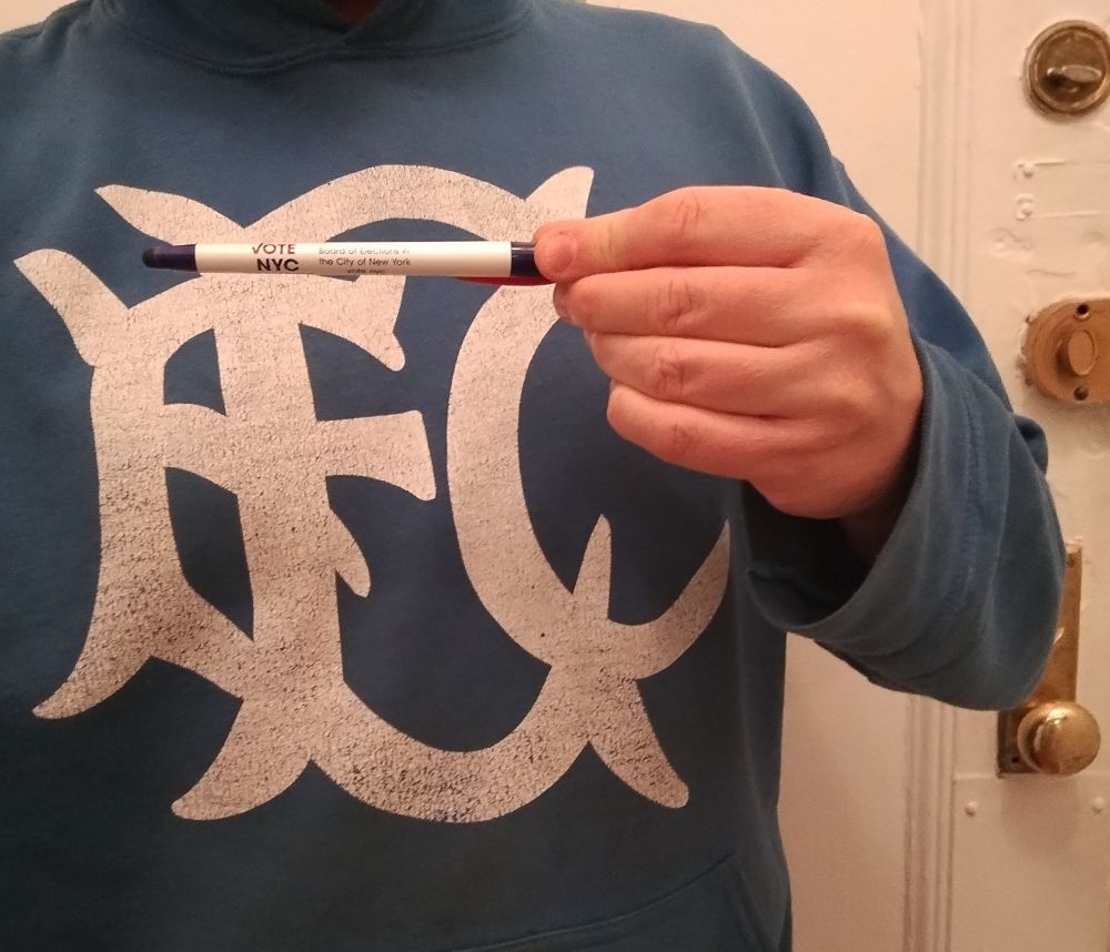 Close up of a.middle aged man holding up a NYC Board of elections pen against an EFC (Everton football club) 