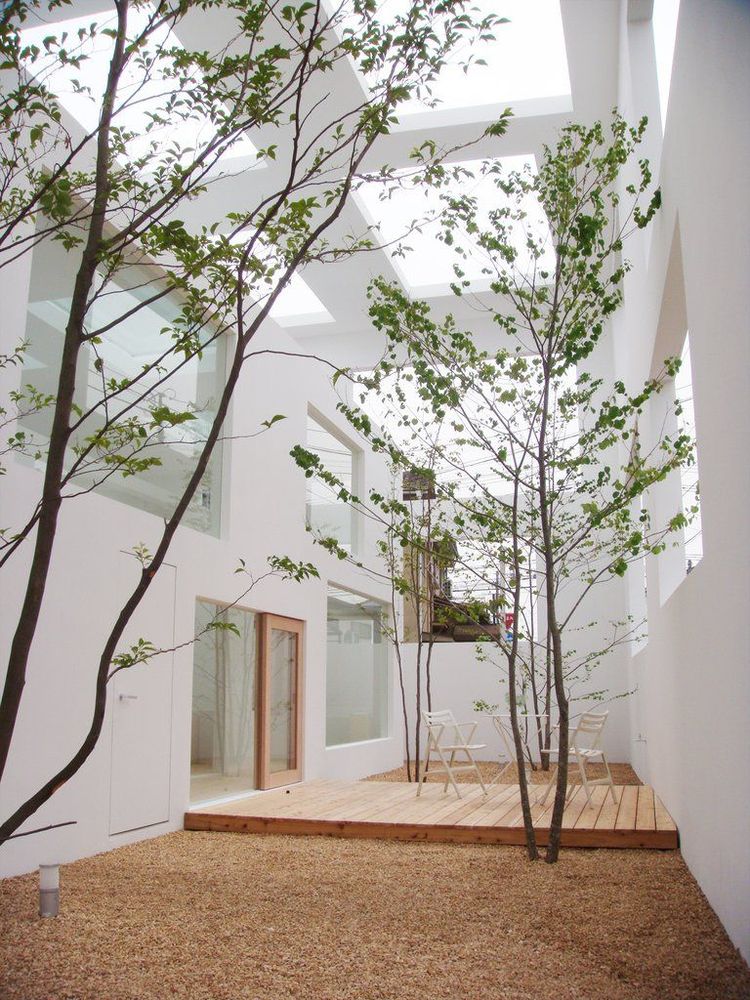 This image shows a minimalist and serene interior courtyard with a modern architectural design. The space features clean white walls with large openings, allowing natural light to flood in. Slender trees with sparse green foliage are planted directly in the gravel-covered ground, creating an organic contrast against the sleek architecture. A small wooden deck with simple white chairs provides a tranquil seating area. The combination of natural elements and modern materials creates a harmonious and calming environment, emphasizing simplicity and connection to nature.