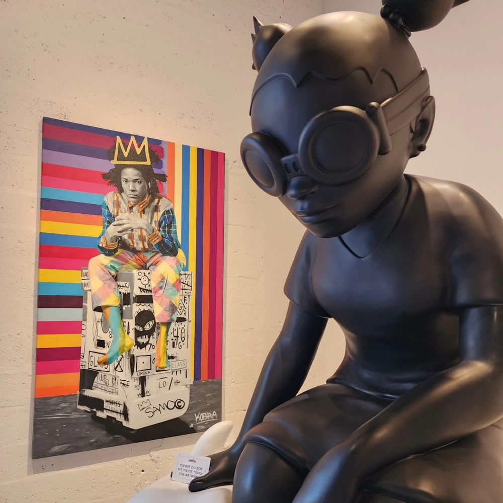 Two artworks: in the foreground a large sculpture of a little girl with steam punk goggles; in the background a painting of Basquiat.