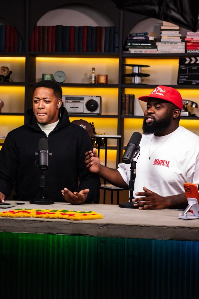 Two podcasters, both men, at a desk with microphones. The man on the left wears a black hoodie and shrugs with his hands open in a questioning gesture. The man on the right, wearing a white t-shirt, red baseball cap, and a beard, speaks emphatically with his hand raised. A colorful yellow, red, and green shaggy rug lies on the desk.