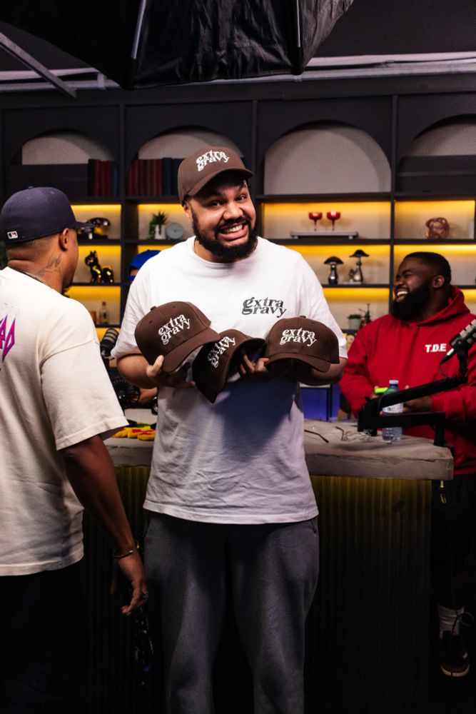 A smiling man proudly holds three brown “extra gravy” caps while wearing a matching shirt and hat. He stands in the Extra Gravy studio with shelves of decor in the background. Two other people are visible nearby, one laughing and another turned slightly away.