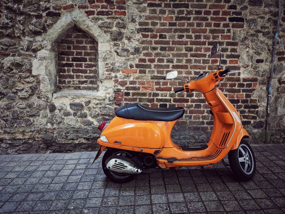 An antique orange Vespa scooter parked in front of a five or six hundred year old church wall located in Aldgate, east London 