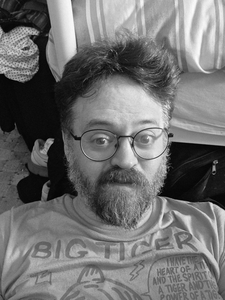 A middle-aged man lying on the floor against a bed post, pulling a silly face.