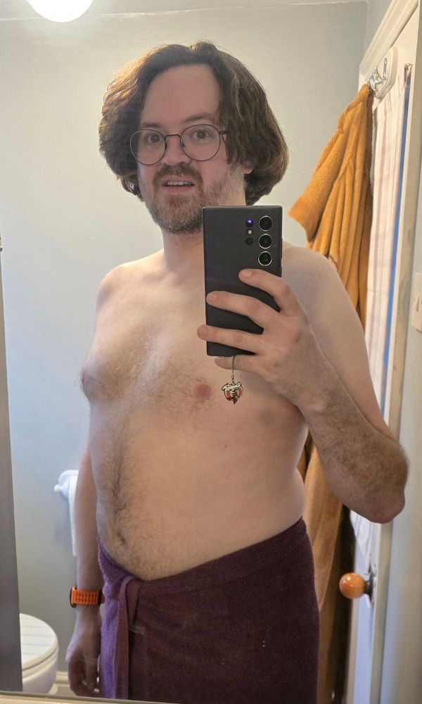 A middle aged man standing at a three quarters angle, taking a mirror selfie in a bathroom wearing nothing but his glasses, a smartwatch and a towel.