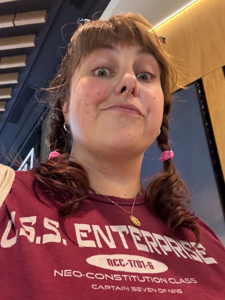 low angle shot of a 30 something women with brown hair in two braids and a burgundy shirt with USS ENTERPRISE NCC-1701-G emblazoned on it