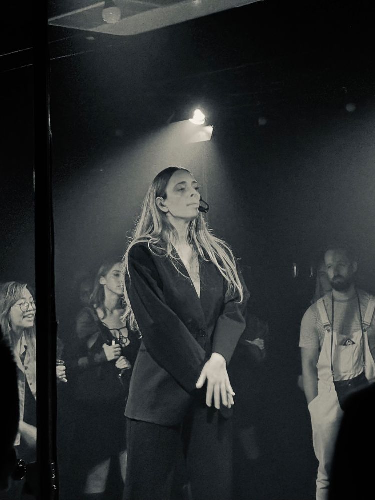 a black and white photo of singer chelsea jade on stage. she is wearing a black suit with long loose blonde hair and a headset. there are people in the crowd behind her