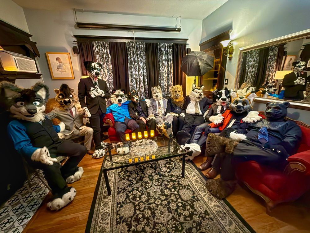 A panoramic photo of a Victorian-style living room filled with different fursuits, each wearing business wear or fancy dress while posing on a red velvet sectional couch.