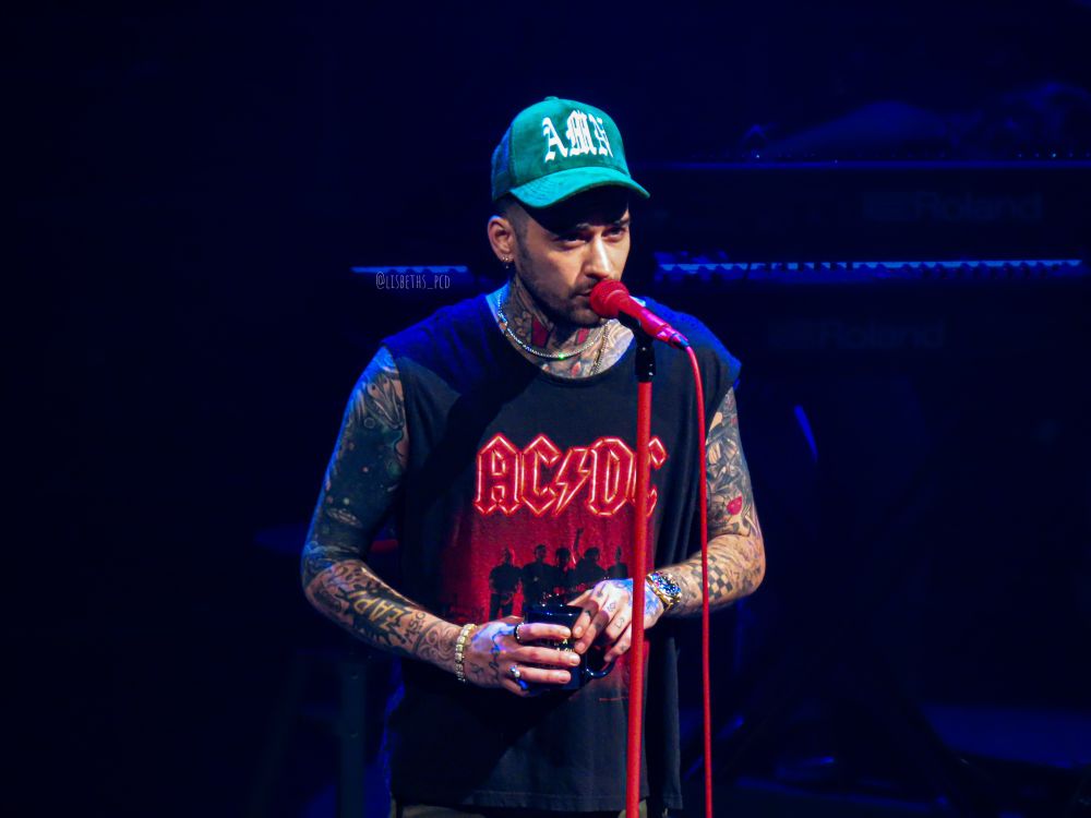 Zayn speaking into a red microphone while he‘s holding a black cup. He is wearing a black shirt with a red AC/DC print and a green cap with the white letters AMN written on it