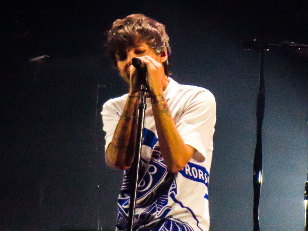 Photo of Louis Tomlinson singing on stage. His eyes are closed, his head is tilted to his right side and he has both hands wrapped around the microphone. He is wearing a white shirt with a blue print on it that shows a knight with a banner and shield riding a horse.