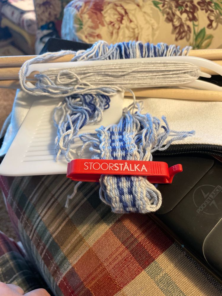 A small section of woven band in light blue and silver with a small stripe of dark blue.  The heddle and shuttle are also visible along with the bright red stoorstalka band lock