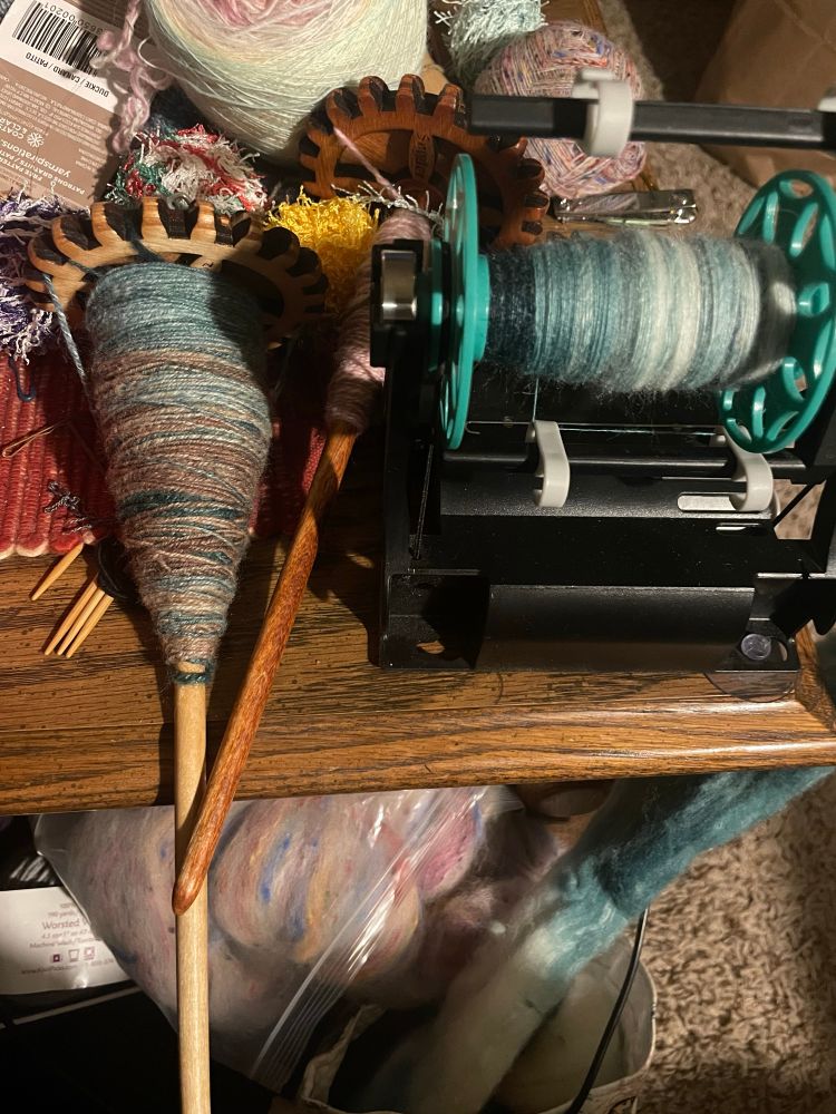 Left, gear spindle with green/white/brown yarn, the spindle is about a third full.  Right, singles of the same fiber on a small electric eel wheel.  Center, pink and white singles being plyed on a spindle, lurking in the back a finished skittles fiber turtle.  Send help, that’s not even all my spinning projects 