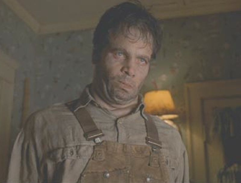 A brown haired man (Vincent D'Onofrio in the film MIB) dressed in a brown pair of dirty overalls and a dirty work shirt makes a twisted face as if drunk or possessed by an alien