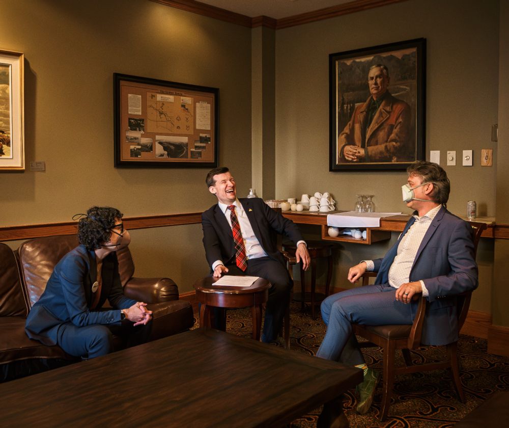 Photo of three people sitting in a room with framed pictures. They are wearing suits and sitting on a sofa and chairs. The middle person is laughing.
