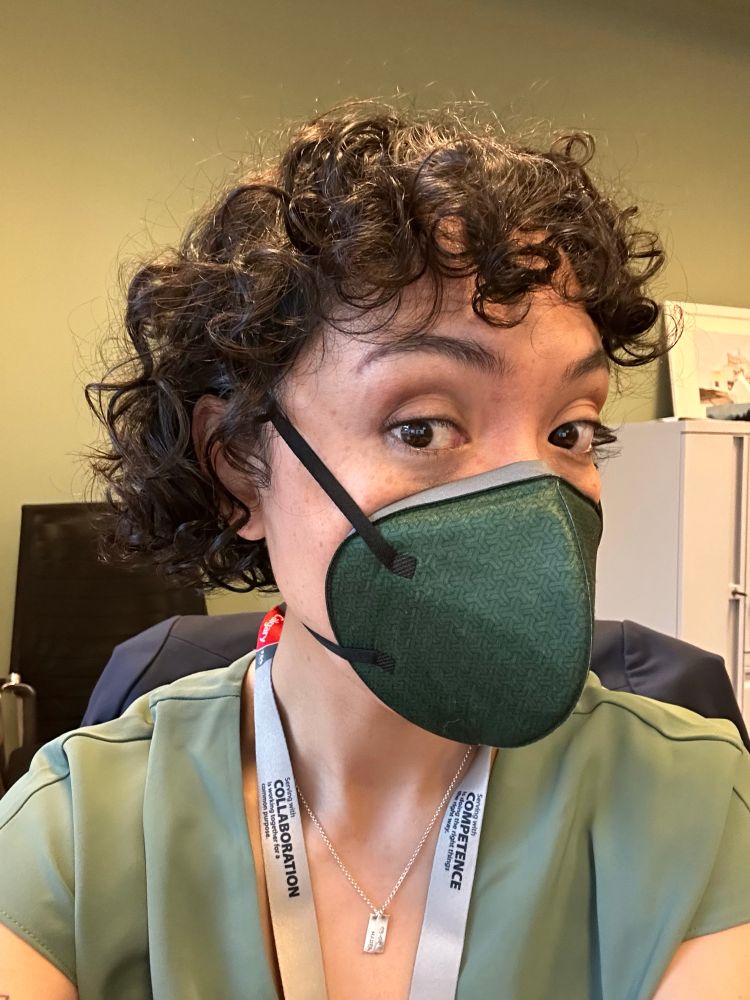 Picture of woman with black curly short hair wearing a green respirator mask, green shirt, sitting in front of a green wall