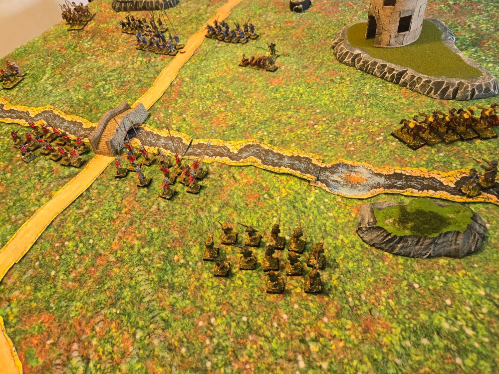 Two 28mm armies face off across a stream and a bridge. A ruined tower stands in the distance.