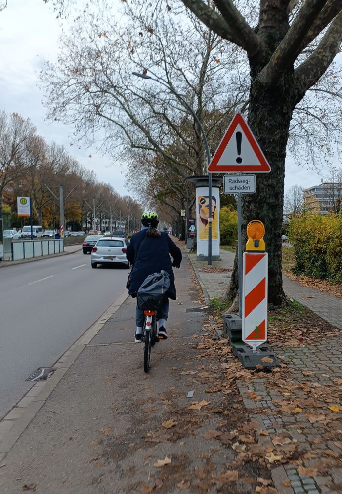 Foto eines Radwegs links von einer Straße. Mittig ist eine Radfahrende zu sehen, rechts davon eine Warnbake und das Schild 