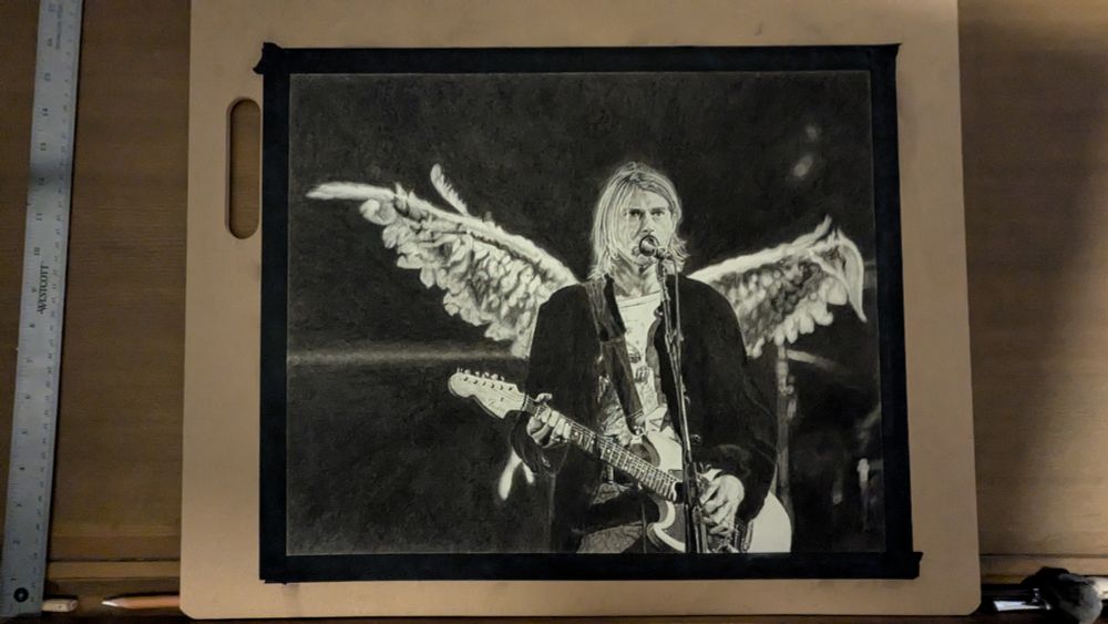 A photo of my finished Kurt Cobain charcoal drawing, but it is still taped to a large drawing board with black artist's tape. The photo has not been cropped and she's the tape, drawing board, and the wooden drawing table where I work, which has a mess of charcoal pencils, charcoal sticks, erasers, and other tools in the tray along the edge of the table.