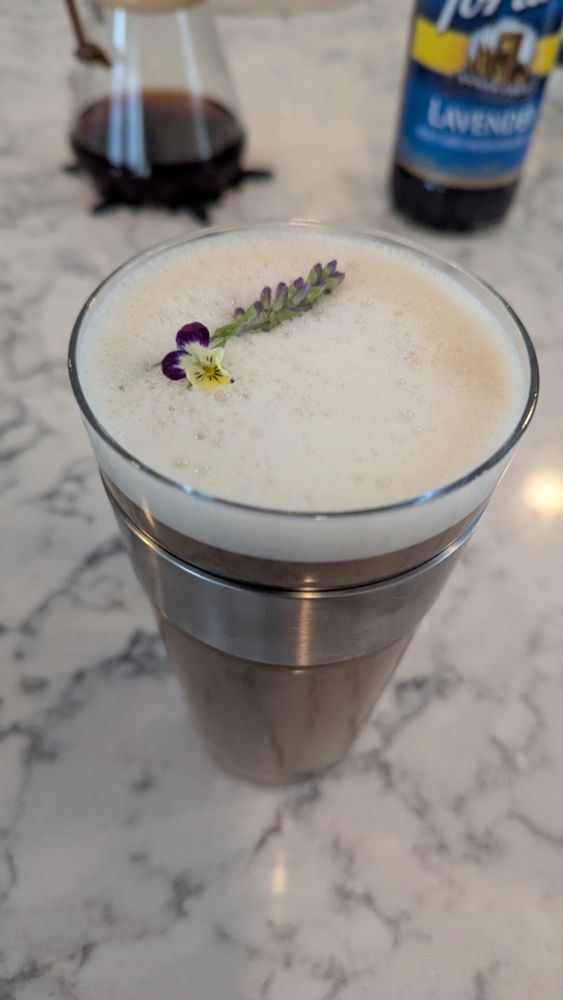 A café au lait in a glass latte mug, topped with oat milk foam and tiny lavender and viola flowers