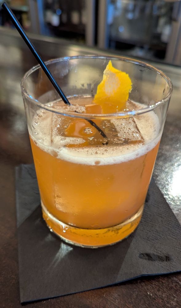 A Paper Plane cocktail (bourbon, aperol, nonino, lemon) in a glass with a large ice cube and an orange peel