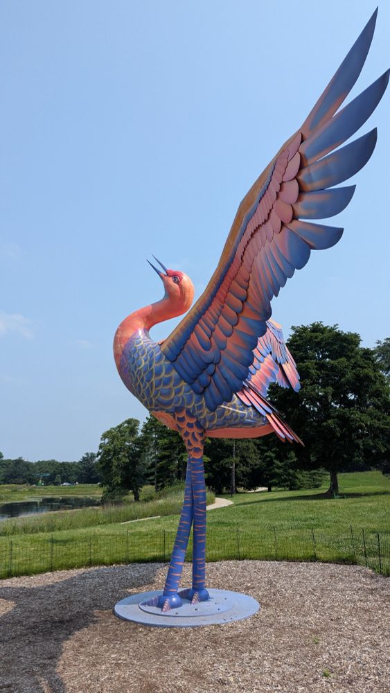 A huge pink and blue sandhill crane sculpture. The bird is standing with its wings spread wide and its head arches slightly back. A pond, walking path, and trees are visible in the background.