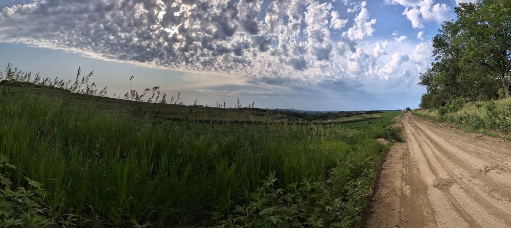 An easy dirt track off to the right, rolling green fields to the left and to the horizon, and a storm brews in the distance with shafts of dark gray rain streaking the sky between shafts of sunlight.