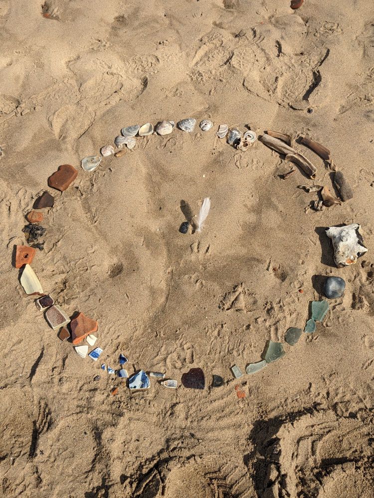 a mandala of flint stones, bones, oyster shells pieces of ceramic (red as well as blue on white) on a sandy beach. In the middle of the mandala a white feather. Photograph, Maria Strutz  
