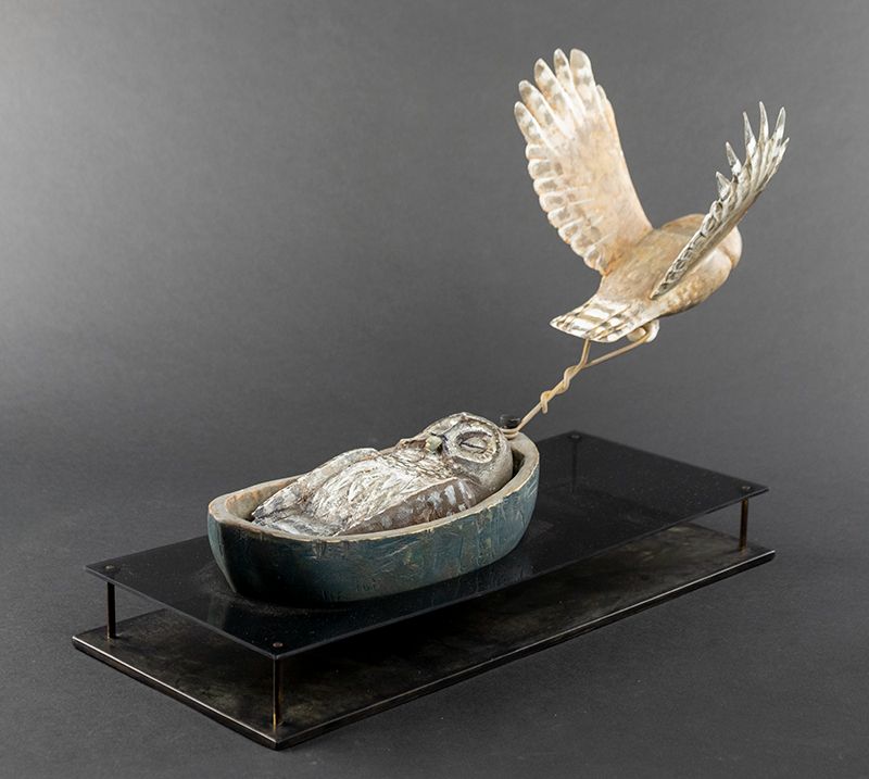 sculpture of an owl flying up and diagonally to the right hand corner, pulling a boat containing an owl with folded wings and closed eyes. Sculpture - Juniper, Pigments, Plexi, Metal Base - by Hib Sabin