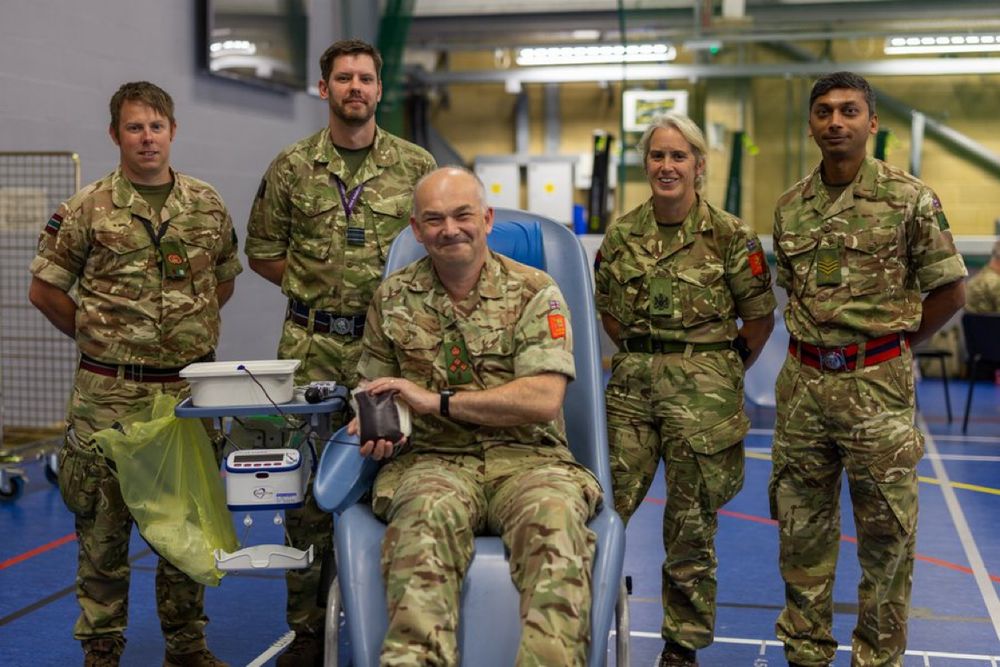 Group of five soldiers in camouflage uniforms inside a gymnasium, one seated donating blood.