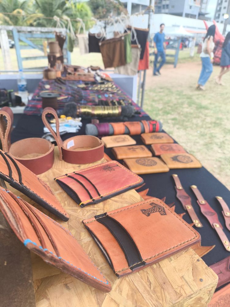 Mesa repleta de itens de couro em primeiro plano. Mais ao fundo tem diversos itens em latão, madeira e bolsas penduradas.