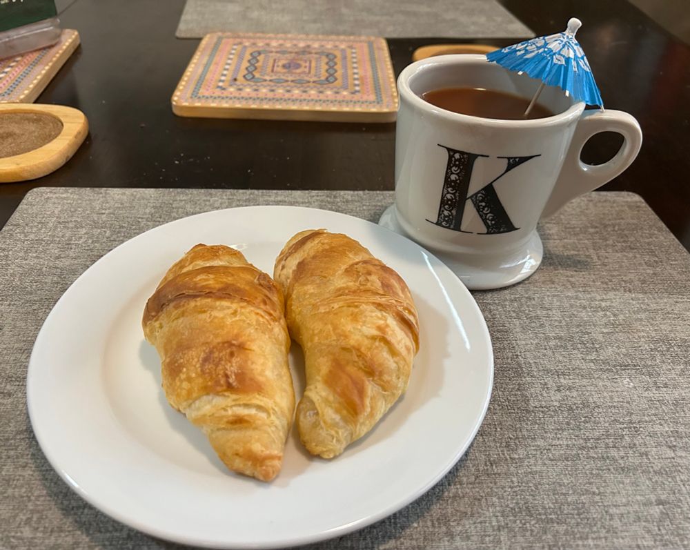 A white plate with two croissants sits on a gray placemat atop a dark wood table. At 1:00 o’clock to the plate is a white mug with a large “K.” The mug is filled with tea, a splash of milk, and — most importantly — topped with a bright blue drink umbrella accented with white flowers. 
