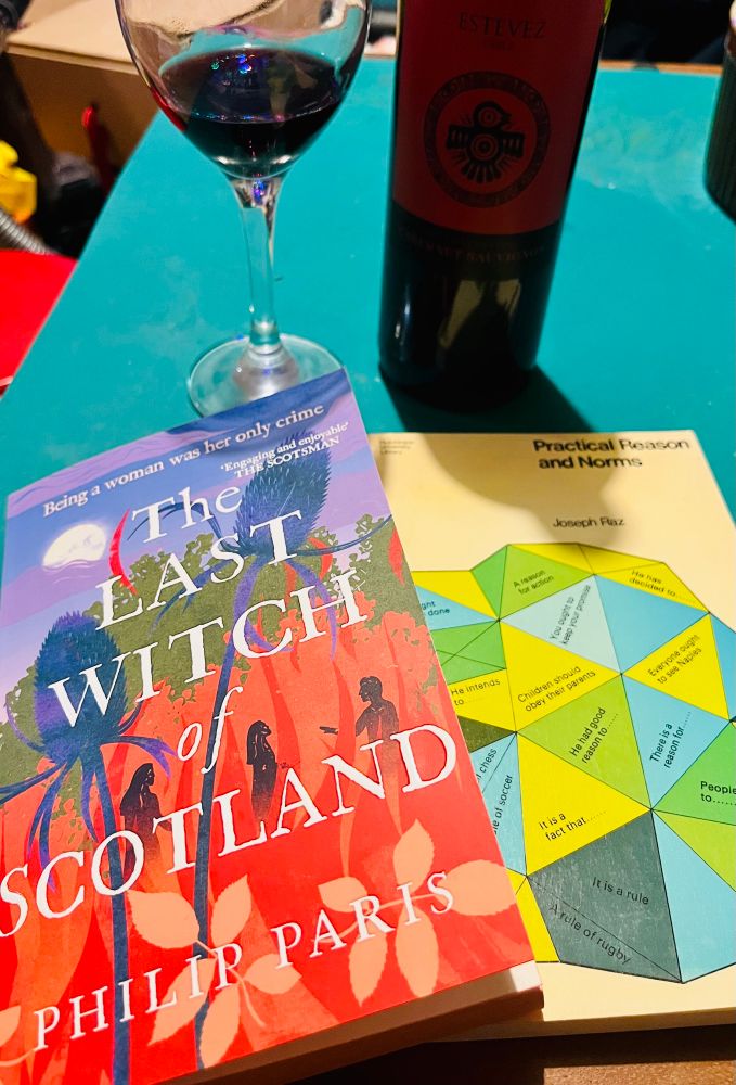 A picture of two books, “The Last Witch of Scotland” by Philip Paris, and “Practical Reason and Norms” by Joseph Raz, with a glass of red wine and a bottle of cabernet sauvignon in the background.
