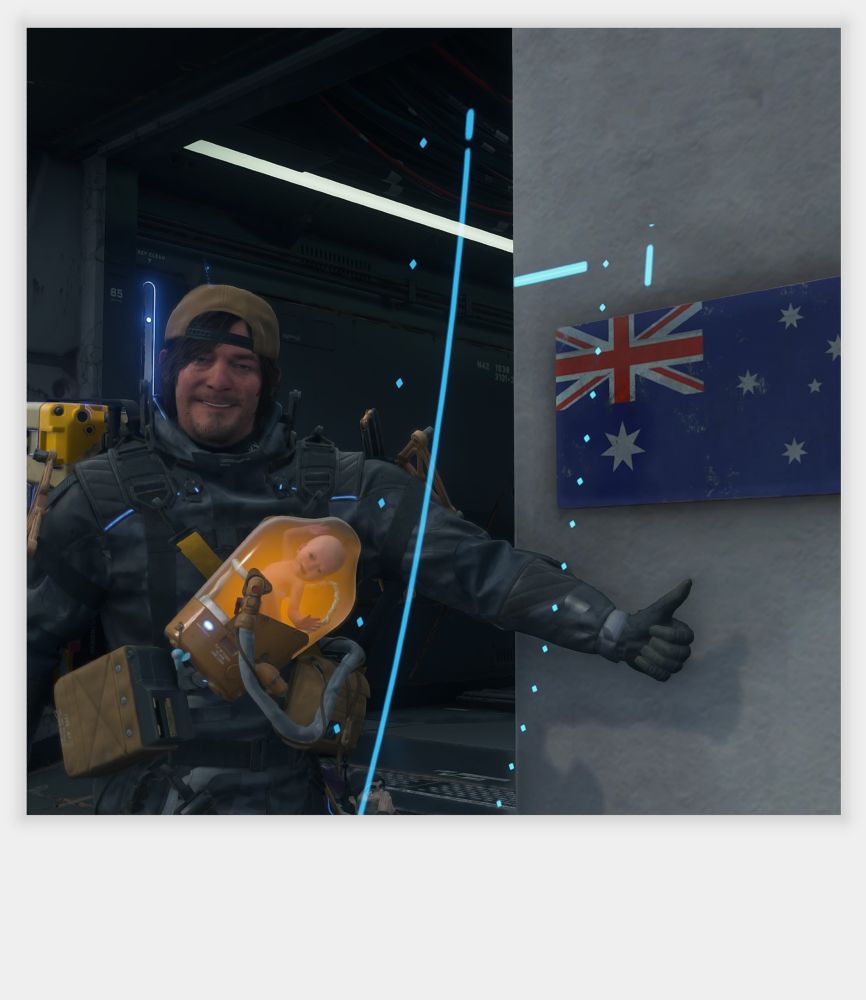 Sam Porter Bridges from Death Stranding 2 On The Beach, standing in front of the first checkpoint once you arrive in Australia. Sam is beside the exterior wall of the facility where the Australian flag is hanging on display. Sam is holding up his thumb next to the flag as he smiles at the camera.