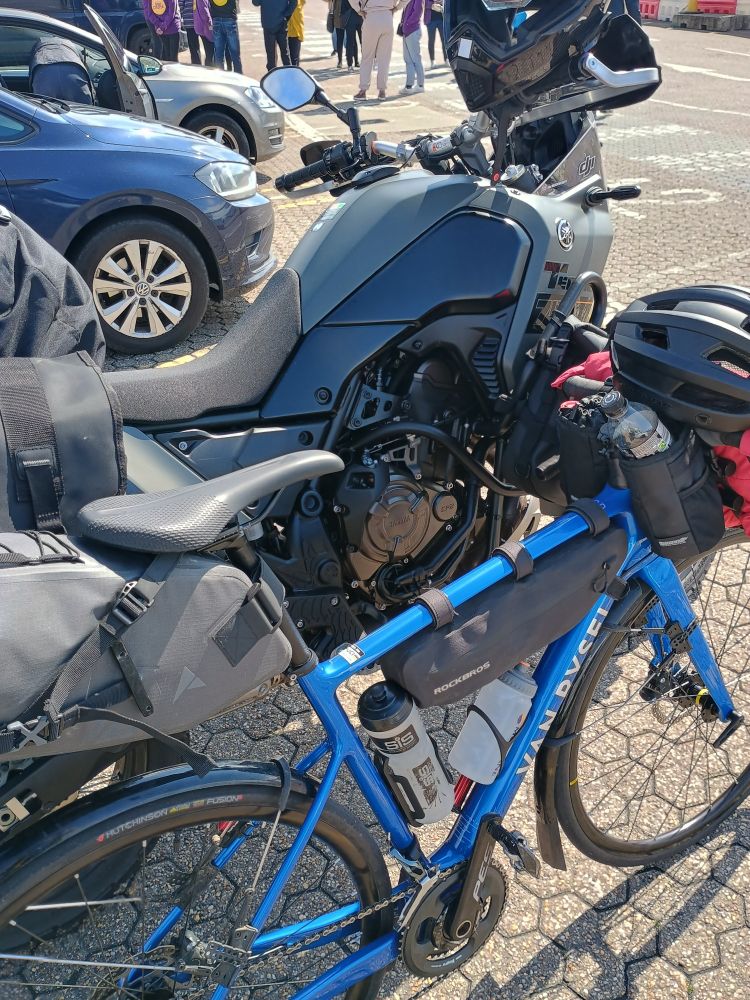 A blue road bike leaning up against a black motorbike. Both bikes have many bags strapped to them!