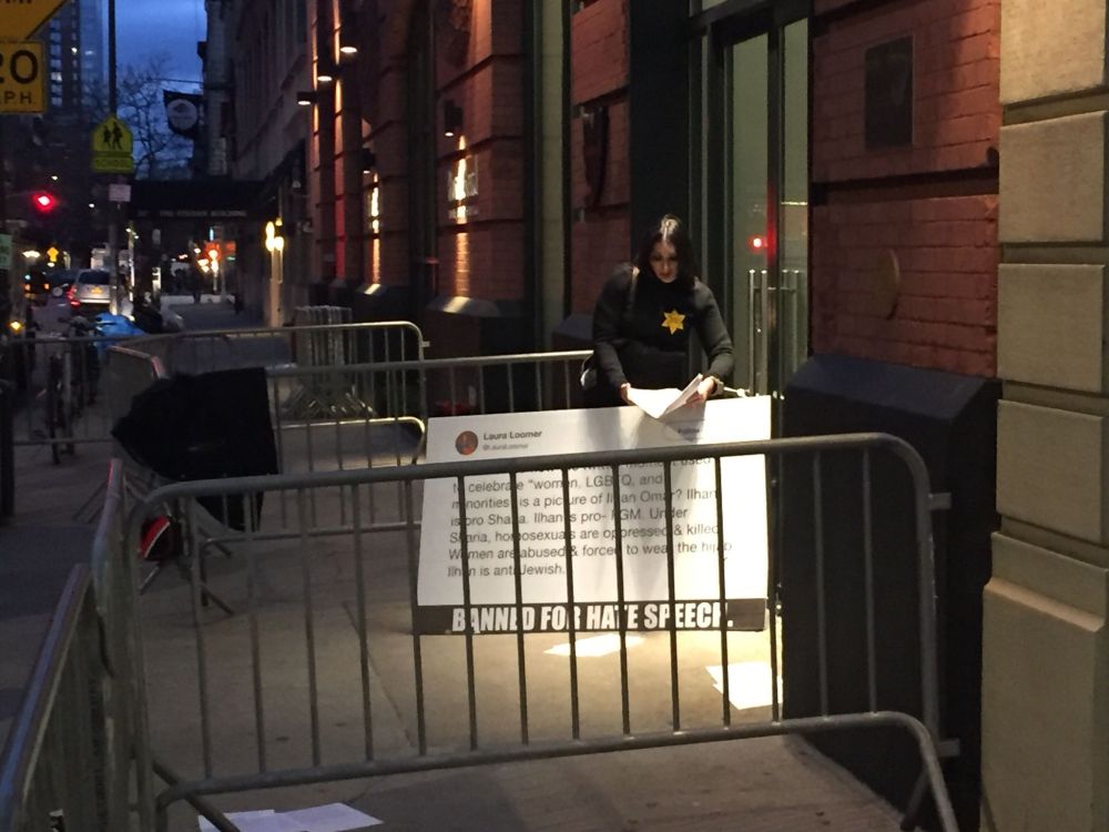 photo shows an empty headed bigot with a bullhorn handcuffed to a glass door, holding a sign on which she’s crying about being banned 