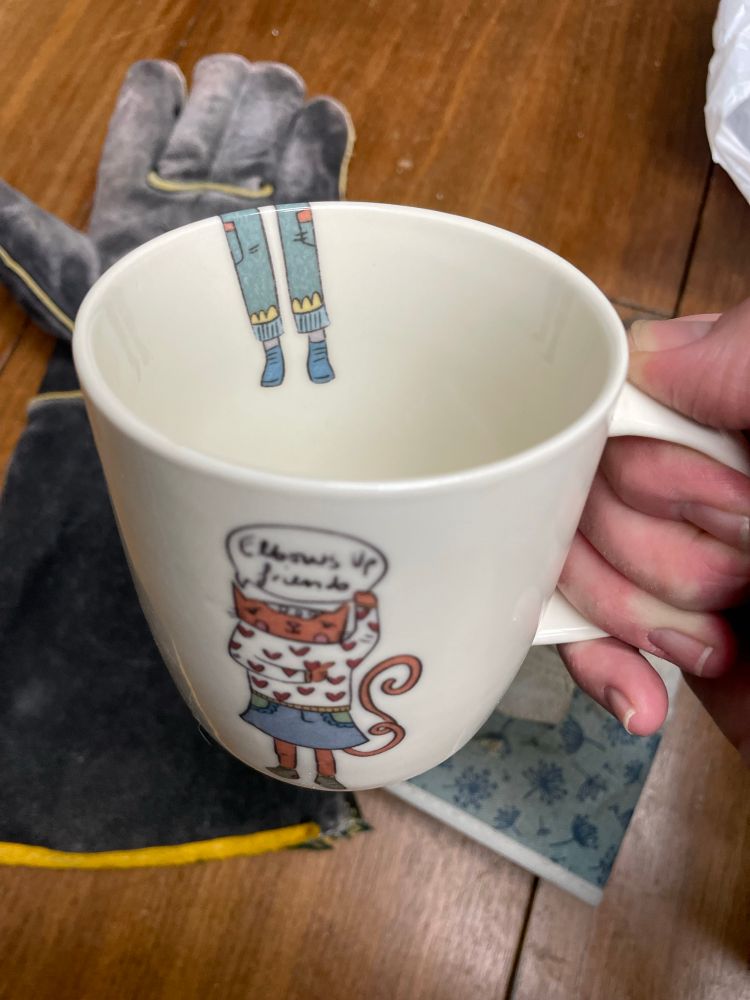 Hand holding a white coffee mug with a drawing of a an orange cat in a sweater covered in red hearts and a jean skirt holds one arm up, other arm point at the elbow of the other arm. A speech bubble says ‘elbows up friends’. You can see two legs in green pants and shoes on the inside of the mug.  