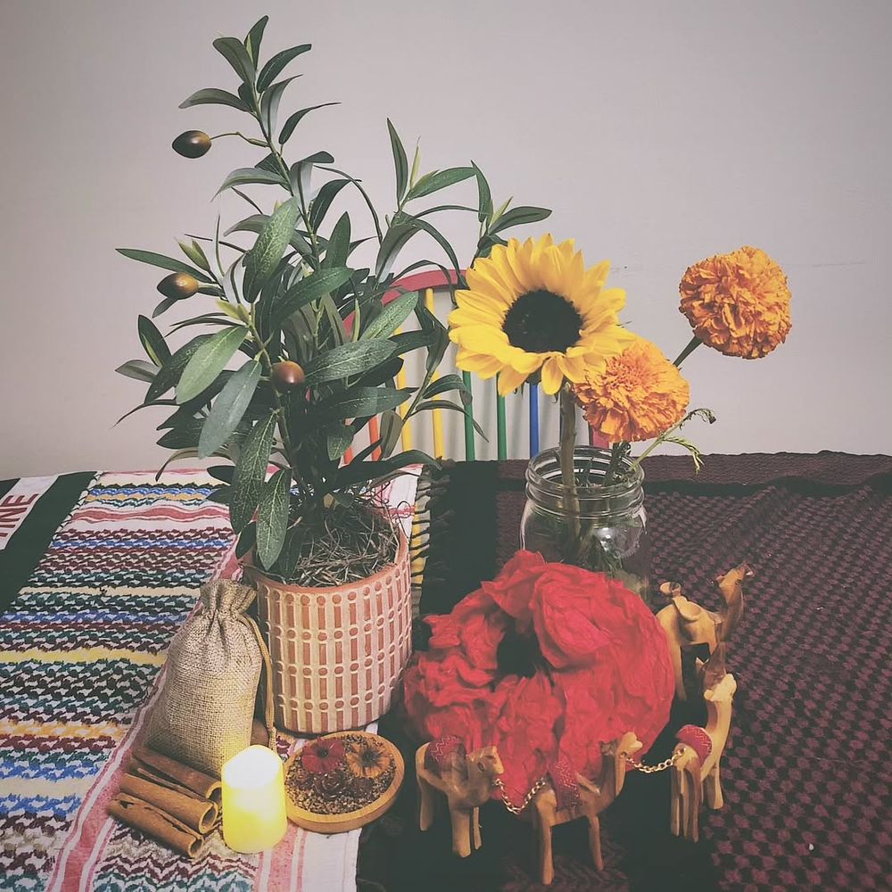 An olive tree plant (not real) next to a mason jar with a sunflower and two marigolds, plus cinnamon sticks, tea light, a poppy made of red tissue paper, and wooden camels, all on top of multi-colored keffiyehs on a table