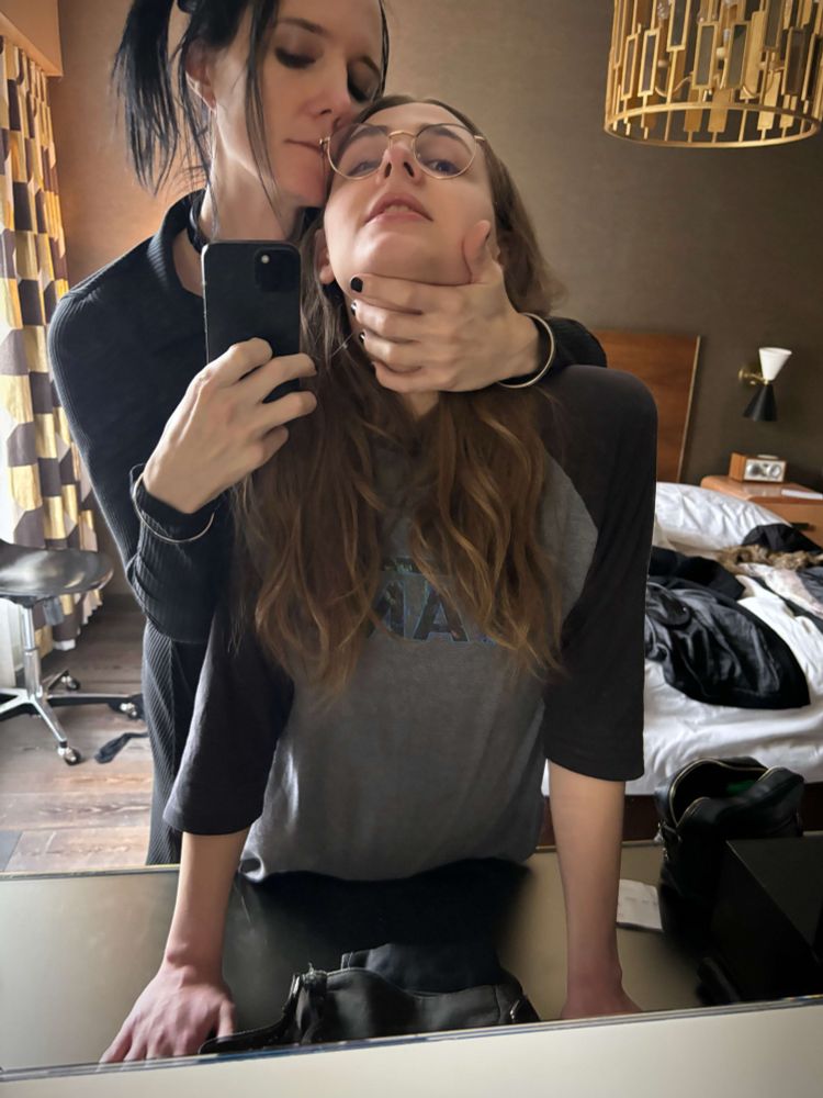 Alice taking a mirror selfie, holding a very good submissive by the throat and bending her over the table.