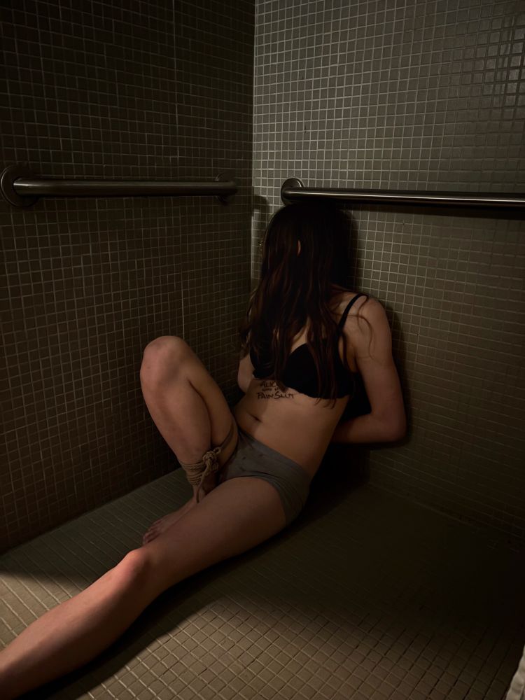A posed shot of me slouched in the bathroom corner after a scene. I am only wearing a bra and panties. My right ankle has been tied to my right thigh, restricting access use of my leg. My arms are held behind my back implying that they’re bound as well. My head is turned away from the camera, with my hair obscuring my face. “Alice’s Good Little Pain Slut” is written on my chest. There is a wet spot on my crotch, implying I either peed myself, or am aroused, those these are actually from my tears T-T.
