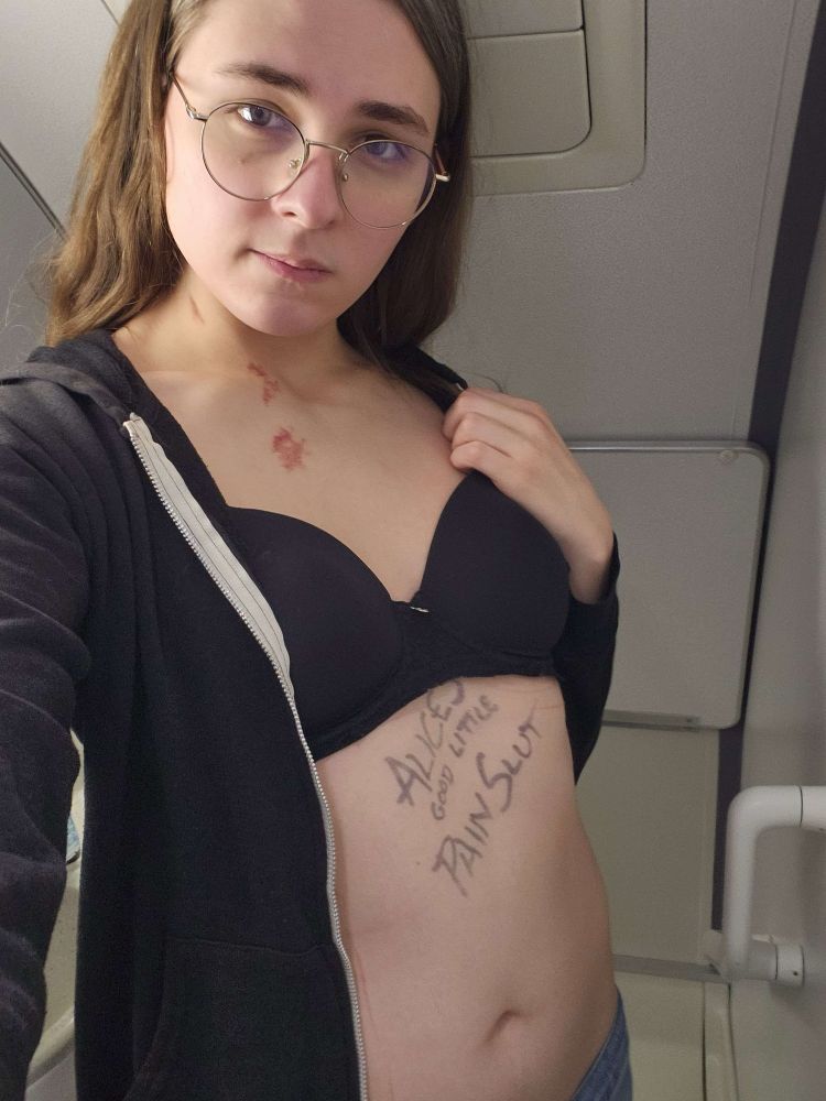 Me taking a selfie in the bathroom of the plane on my flight home from visiting Alice. I have two hickies around my collarbone. It says “Alice’s Good Little Pain Slut” on my chest in black marker.