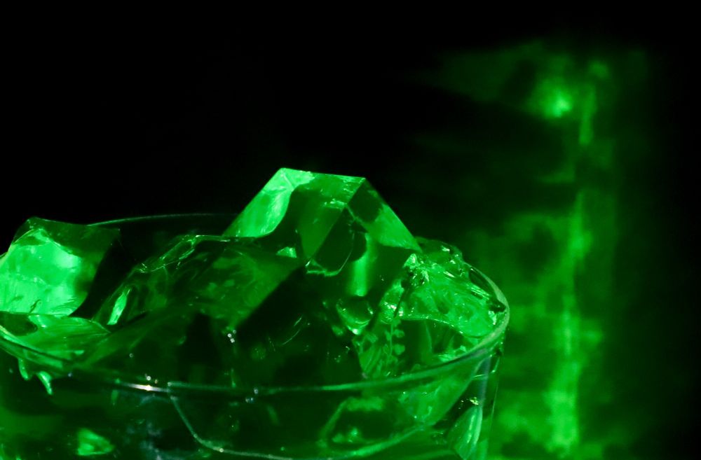Da Foto zeigt Würfel grüner Götterspeise in einem Glas. Der Hintergrund ist schwarz. Das seitlich in das Bild fallende Licht zaubert grüne Reflexionen auf den Hintergrund. 