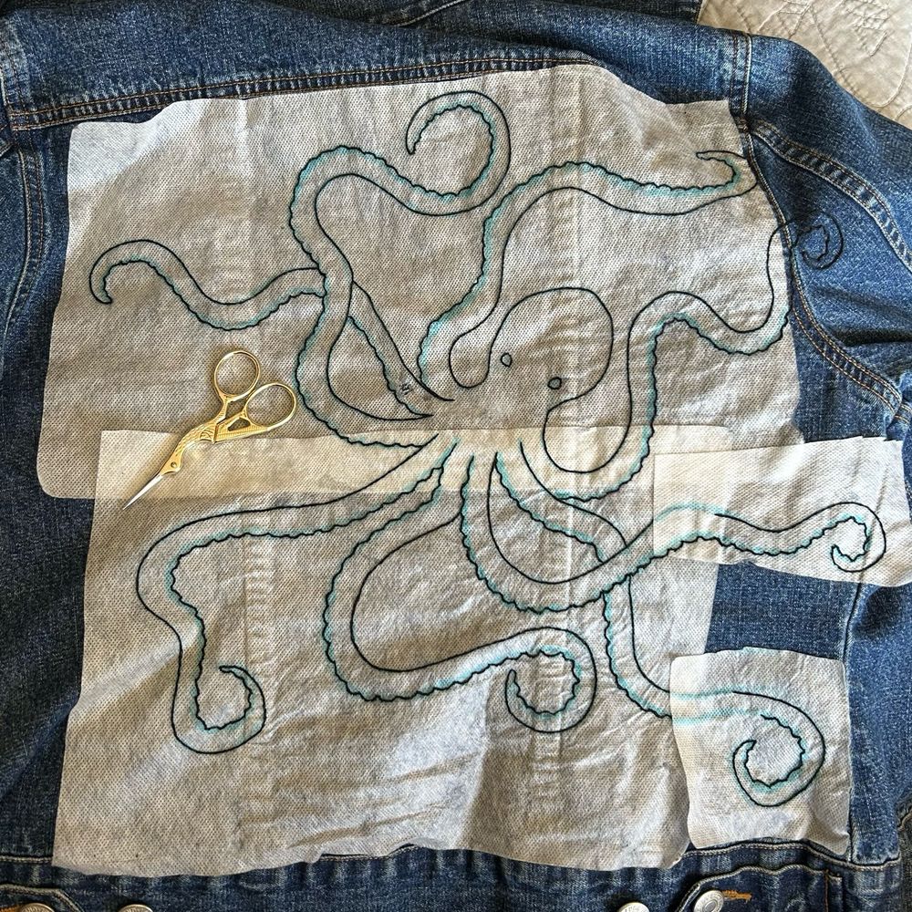 A denim jacket embroidered with a black outline of an octopus with white interface still attached 