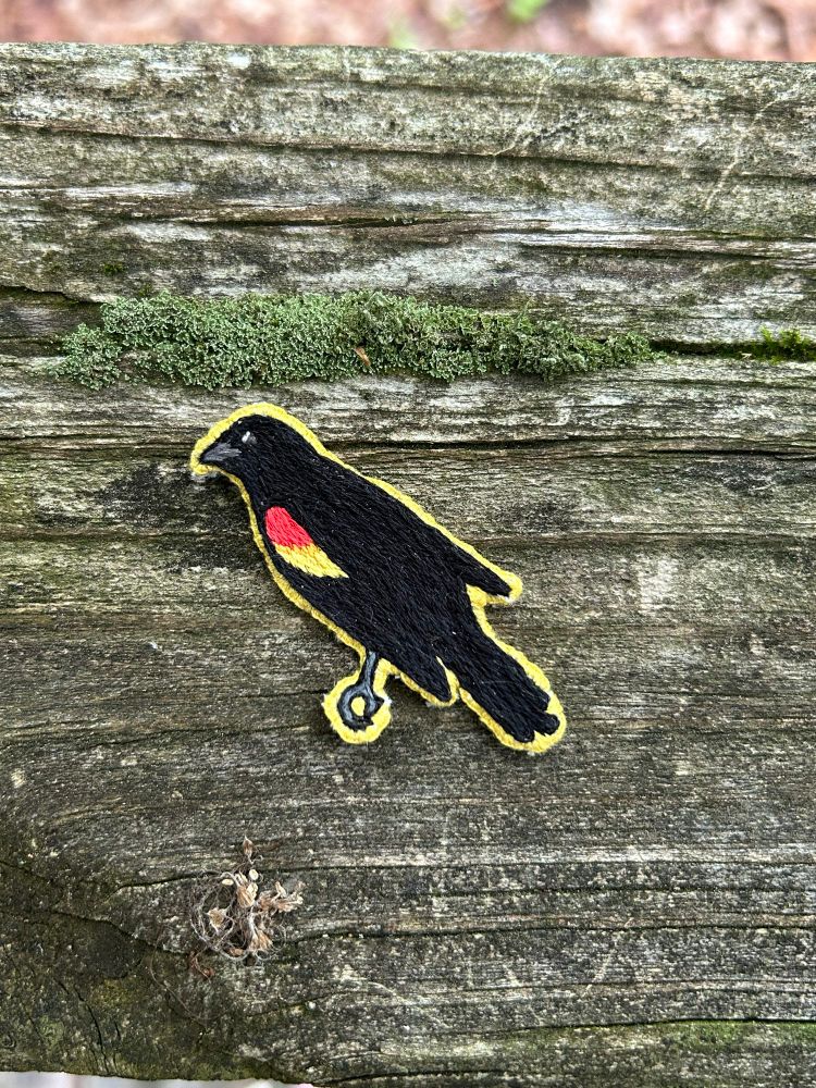 A hand embroidered red-winged blackbird patch. The patches is finished with yellow thread and is sitting on a backdrop of wood and moss. 