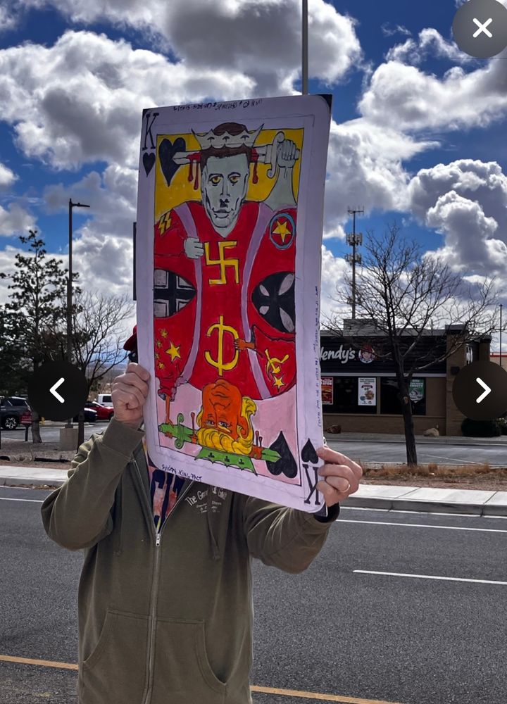 Protester in Bernalillo holds a vibrant poster in the style of a King of Hearts playing card. The poster is the color of a coral snake: scarlet, black, and yellow. Musk and Trump are the kings. Musk is the top. Trump is the bottom. The Musk king has a pallid face and yellow eyes, a Hitler mustache, and a swastika. He seems to be cutting his own head with his own sword. The Trump king’s face is deeply flushed and saturated, like a Flamin’ Hot Cheeto. He has tiny, fragile hands. He wears the hammer and sickle on his scarlet robe.