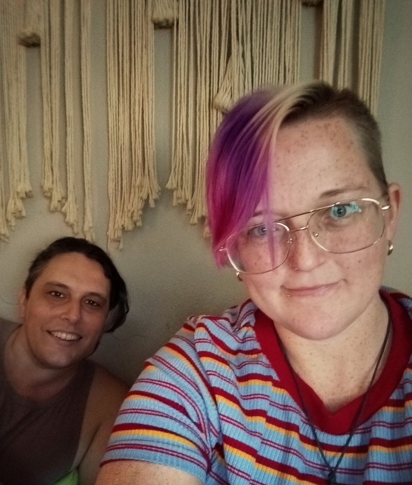 A selfie showing Stevie with pink and purple hair, gold aviator glasses, and a red/yellow/blue striped shirt next to their wife Gwen who has her dark brown hair pulled back in a ponytail and is wearing a pale purple tank top. Both are smiling.