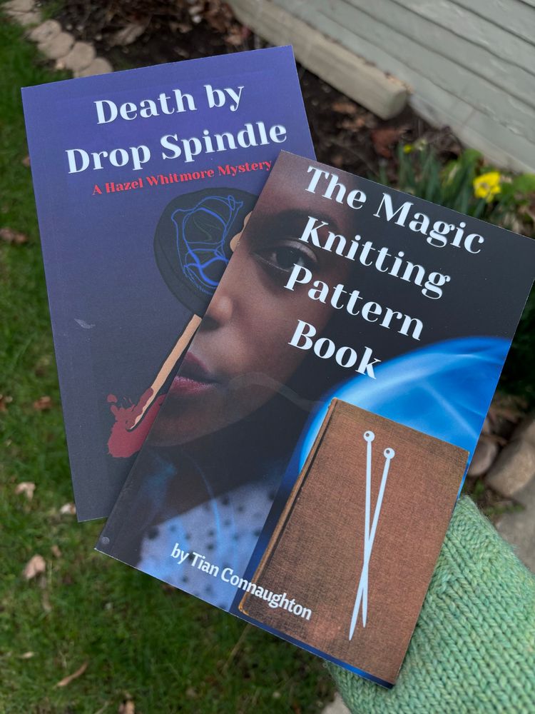 An arm in a mint green sweater holds 2 books: The Magic Knitting Pattern and Death by Drop Spindle, by Tian Connaughton. In the background a few yellow narcissus are starting to bloom.